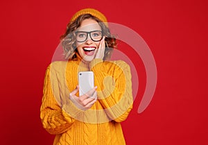 Delighted young female with smartphone touches   cheek  and looks at the camera laughing on a bright red background