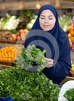 Delighted young Muslim woman purchaser choosing cilantro in grocery store