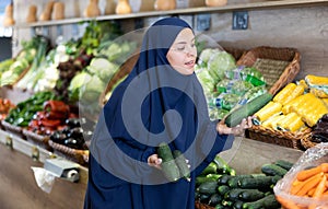 Delighted young Muslim woman purchaser choosing cucumber in grocery store