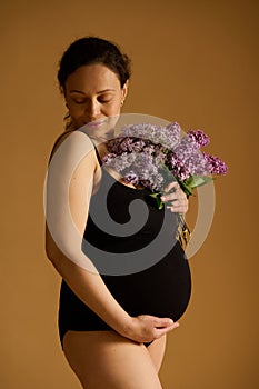 Delightful pregnant woman in black bodysuit, posing with purple lilacs on beige background, enjoying her pregnancy time