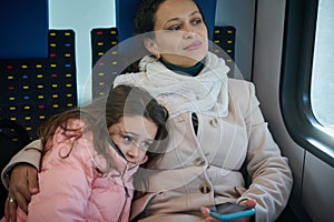 Delightful young woman, happy mother hugging her little daughter, travelling together on a comfortable urban train