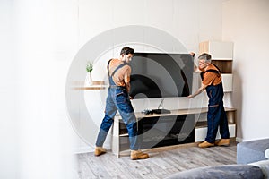 Delivered on time. Two handymen, workers in uniform hanging, installing tv television on the wall indoors. Repair and