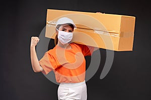 Delivery asian woman or courier wearing an orange masked uniform holds a cardboard box and act strong in the coronavirus concept