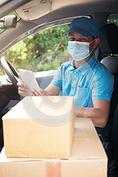 Delivery man driver wearing protective face mask holding digital tablet looking at camera with blurred cardboard boxes on van seat