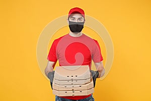 Delivery man employee in red cap t-shirt uniform black mask gloves give food order pizza boxes isolated on yellow