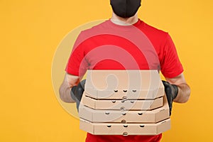 Delivery man employee in red cap t-shirt uniform black mask gloves give food order pizza boxes isolated on yellow