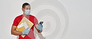 Delivery man in face mask and protective gloves holding parcels, letters and using smartphone with mobile banking