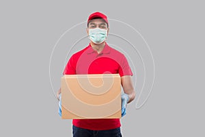 Delivery Man Holding Box in Hands Wearing Medical Mask and Gloves Isolated. Red Tshirt Indian Delivery Boy Watching Side. Home