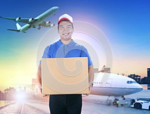 delivery man holding card box toothy smiling face against shipping port and cargo plane flying background