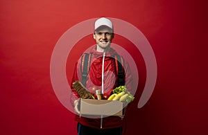 Delivery man in red uniform holds cardboard paper box with food isolated on white background, studio portrait. Service