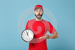 Delivery man in red workwear hold clock isolated on blue wall background studio portrait. Professional male employee in