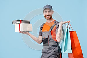 Delivery man in uniform holding shopping bags and gift boxes on blue studio background