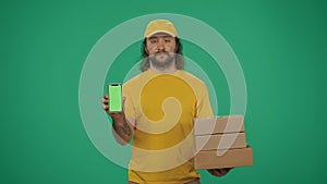 Delivery man in uniform holding smartphone and stack of postal boxes. Isolated on green background. Advertising area