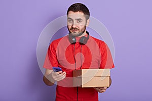 Delivery man using mobile phone for finding exact delivery address while holding package in other hand, posing isolated over lilac