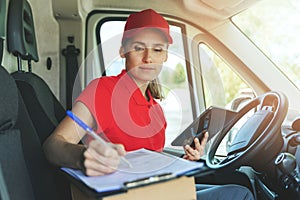 Delivery person in red uniform sitting in van and writing documents on clipboard