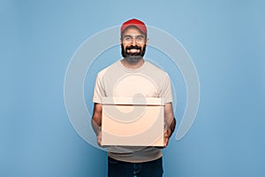 Delivery service concept. Cheerful indian mailman holding cardboard box and smiling at camera, blue background