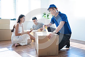 Delivery service worker man in blue uniform help happy couple customer packing parcel with tape in customer home.Woman with tablet