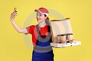 Delivery woman holding coffee and pizza boxes, taking selfie or broadcasting livestream.