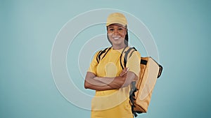 Delivery woman in uniform with portable backpack refrigerator, posing at the camera with crossed hands. Isolated on blue