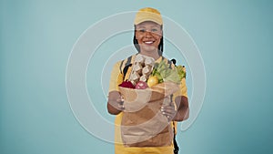 Delivery woman in uniform with portable refrigerator giving paper bag with grocery products. Isolated on blue background