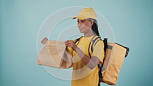 Delivery woman in uniform with portable refrigerator holding paper bag giving it to client. Isolated on blue background.