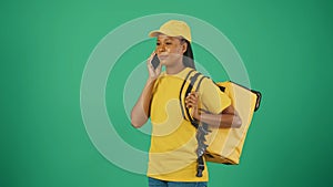 Delivery woman in uniform with smartphone and portable refrigerator, calling her client. Isolated on green background.