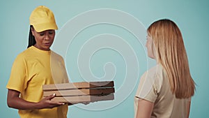 Delivery woman in yellow uniform giving female customer order stack of pizza boxes. Isolated on blue background.