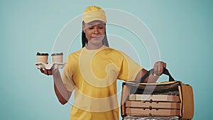 Delivery woman in yellow uniform holding coffee cups holder and refrigerator backpack with pizza boxes. Isolated on blue