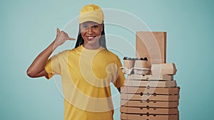 Delivery woman in yellow uniform pointing at coffee cups and pizza boxes, shows calling gesture. Isolated on blue