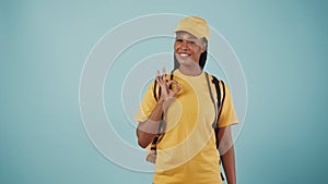 Delivery woman in yellow uniform with portable backpack refrigerator shows ok sign. Isolated on blue background.