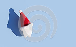 dental christmas, new year concept. toy tooth in red santa hat with hard shadows on blue background.