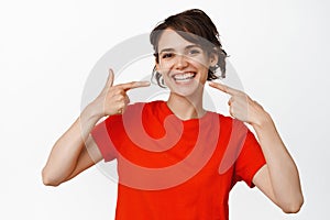 Dental clinic, stomatology concept. Smiling brunette woman pointing at her perfect white teeth, looking satisfied, white