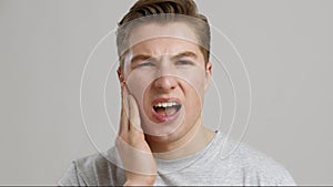 Dental pain. Young man suffering from acute toothache, rubbing his sore cheek, grey studio background