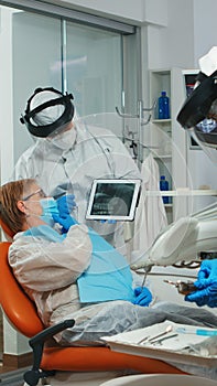 Dentist in coverall showing on tablet dental x-ray reviewing it