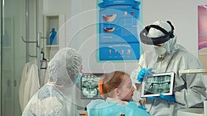 Dentist in protective equipment showing on tablet dental x-ray of child