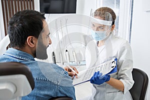 Dentist in protective mask talking her male patient