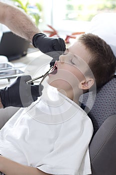Dentist`s hands in black rubber gloves hold tongs for pulling teeth. The boy with his eyes closed sits calmly in the chair wit
