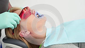 Dentist's hands puts protective red glasses on female patient's face before UV teeth whitening. Blonde woman