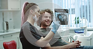 Dentist shows to his patient in a light dental clinic room teeth X-ray , and explain to him some details of his oral