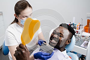 Dentistry. Dentist showing teeth color shades guide for tooth whitening for african patient in dental clinic. African is