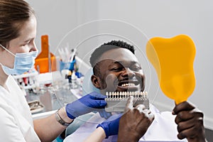 Dentistry. Doctor checking teeth color matching samples in dental clinic. Teeth whitening of african man. Dentist