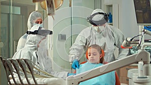 Dentistry nurse in coverall putting dental bib to child before examination