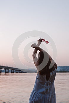 a depersonalized young woman with her back in a white sundress with her arms crossed over her head after sunset at the photo