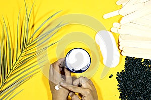 Depilatory pearly blue solid wax beans and wooden stick, cotton pads and woman making hand cream box and palm leaf on yellow backg