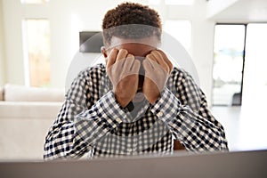 Depressed African American  teenage boy with head in hands using a laptop computer at home, front view, close up