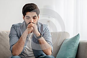 Depressed arab man having problems, thinking while sitting on couch at home, empty space