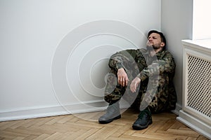 Depressed army man in uniform sitting in a corner of an empty room. Place for your poster on the wall