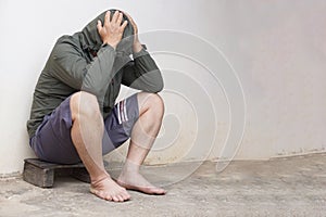 Depressed Asian man wears hoodie shirt, shorts, bare feet, sits lean old wall on pavement. Concept, Life problems