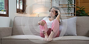 Depressed Asian senior woman sit on sofa looking away by the window. Loneliness elderly female living alone at home with