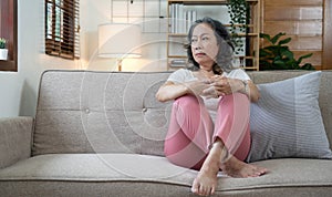 Depressed Asian senior woman sit on sofa looking away by the window. Loneliness elderly female living alone at home with